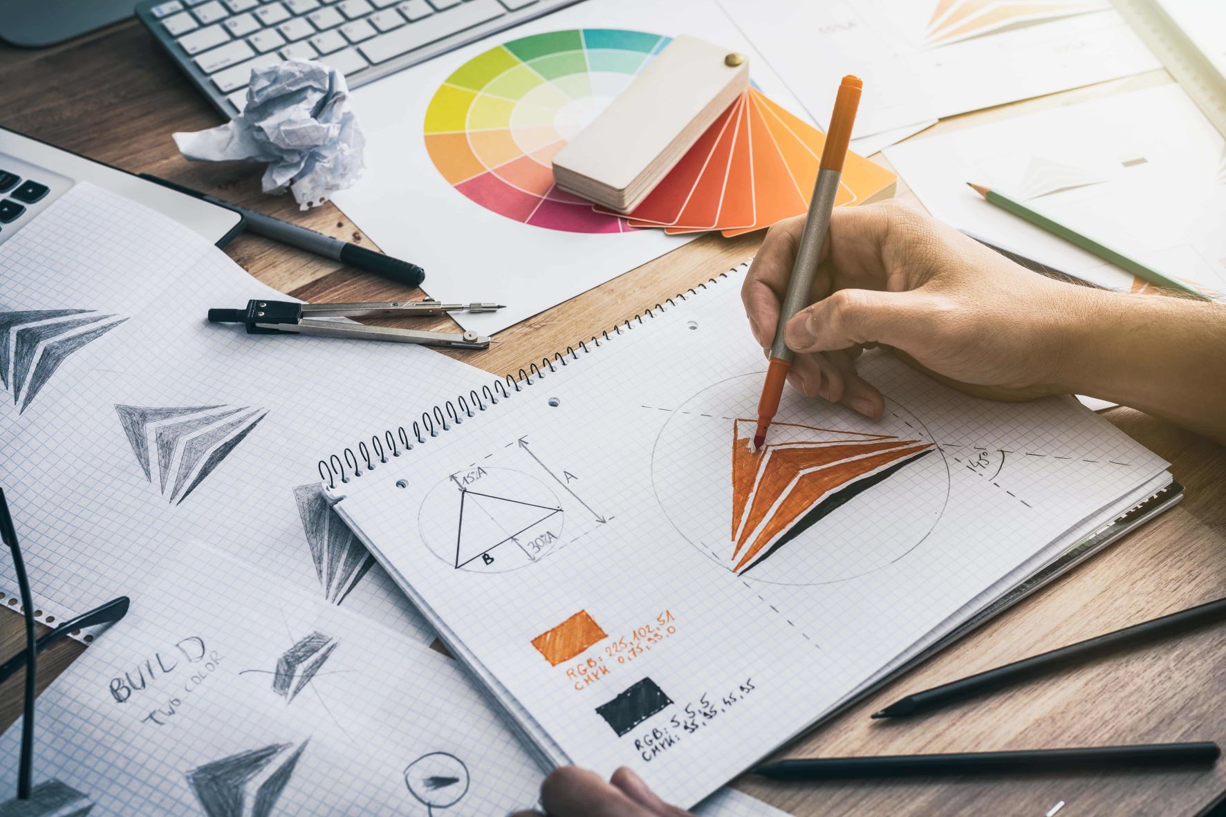 Una persona dibuja formas geométricas en un cuaderno con un lápiz, rodeada de muestras de color, papel y herramientas de dibujo sobre un escritorio: un enfoque artístico de la marca y la consultoría empresarial que transforma las ideas en narrativas visuales.